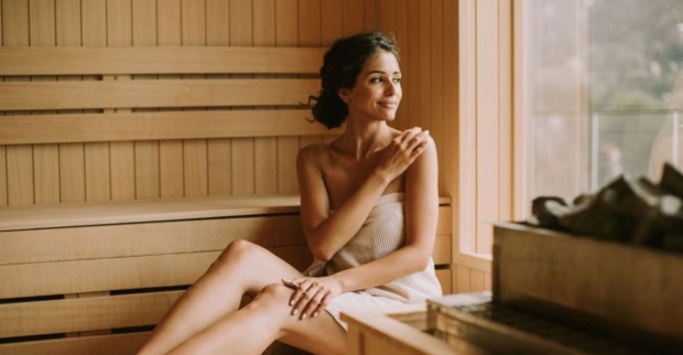 Eine Frau sitzt mit einem Handtuch bedeckt in einer gemütlichen Sauna aus Holz und schaut glücklich aus dem Fenster neben ihr.