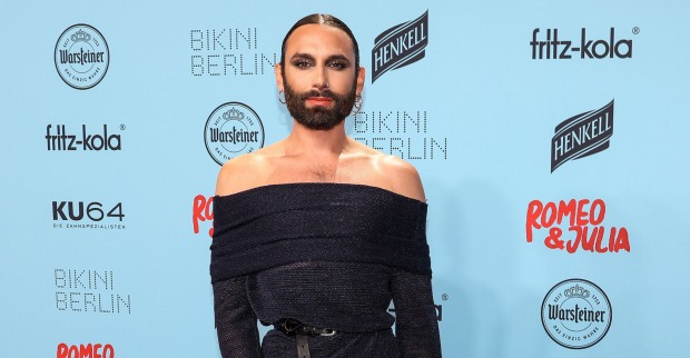 Conchita Wurst mit einem engen schwarzen Kleid und gegelten Haaren vor einer hellblauen Fotowand.