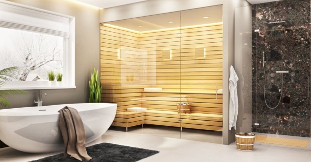 Ein Badezimmer mit einer weißen freistehenden Badewanne, einer Dusche mit dunklen Wandfliesen und eine moderne Sauna im Holzstil, die mittels Glasfront vom restlichen Raum abgetrennt ist.