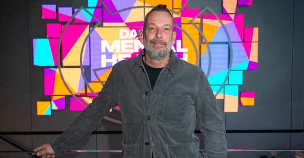 Robert Kratky trägt eine dunkle Weste und steht in einem Studio mit bunten Hintergrund.