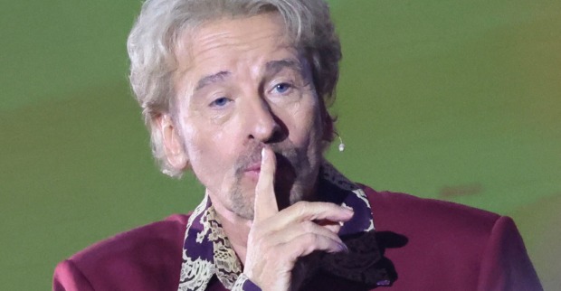 Thomas Gottschalk hält sich den Zeigefinger schweigend vor den Mund. Er trägt ein weinrotes Sakko.