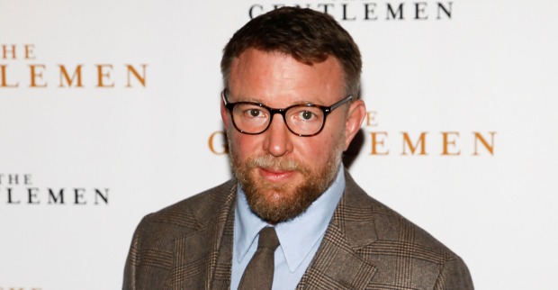 Guy Ritchie vor einer Fotoleinwand mit einem beigen Sakko.