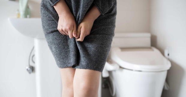 Eine Frau muss dringend auf die Toilette. Sie zwickt die Beine zusammen. Im Hintergrund die rettende WC-Schüssel.