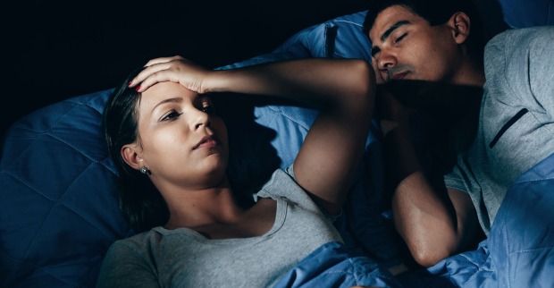 Paar liegt zusammen in einem Bett, ein Partner wach, der andere schläft daneben.