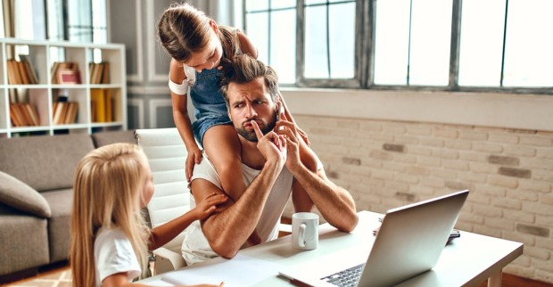 Ein junger Vater sitzt an seinem Schreibtisch vor einem Laptop, rechts neben ihm ein kleines blondes Mädchen, das mit Buntstiften malt und auf seinen Schultern sitzt ein kleines braunhaariges Mädchen | Credit: iStock.com/Valerii Apetroaiei