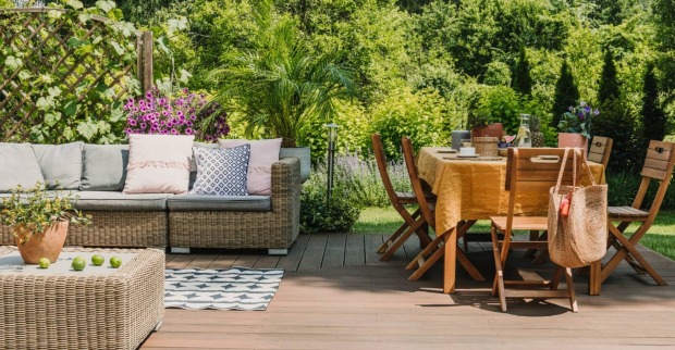 Blick in einen Garten mit Holztisch und Sesseln. Im Hintergrund ein Rattansofa.