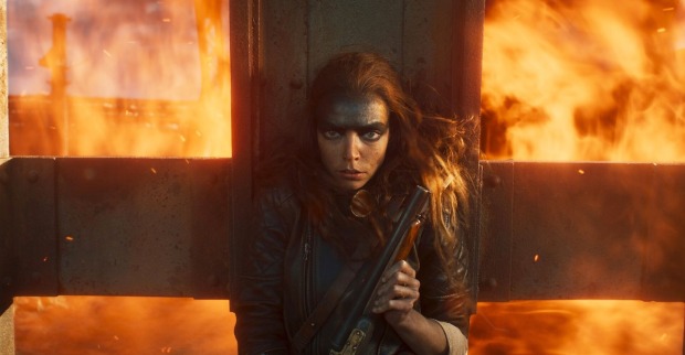 Eine Schauspielerin vom Film "Furiosa: A Mad Max Saga" mit viel schwarzer Schimnke. Hinter ihr sind Flammen. Sie trägt eine Waffe.