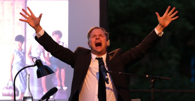 Philipp Hochmair mit einem schwarzen Anzug und einer schwarzen Krawatte auf der Bühne.