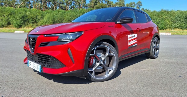 Ein roter Alfa Junior steht auf einem asphaltierten Parkplatz. das Auto glänzt in der Sonne, im Hintergrund sind Föhren und andere Bäume zu erkennen.