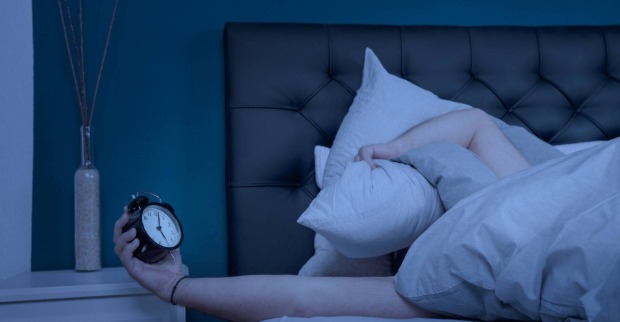 Ein Mann, der in einem dunklen Schlafzimmer mit einer Hand einen Polster auf sein Gesicht drückt, während er in der anderen einen Wecker hält