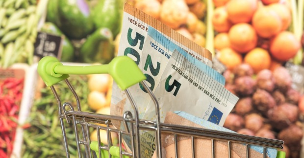 Euro-Geldscheine in einen Einkaufswagen. Im Hintergrund ein Gemüseregal.