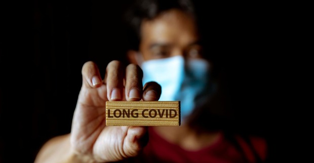 Mann trägt Corona-Maske und hält Holzstück mit Aufschrift "Long Covid" in die Kamera