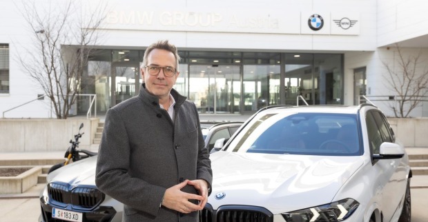 Alexander Bamberger, trägt eine Brille und ist mit einem grauen Mantel bekleidet. Der BMW-Manager steht vor zwei weißen BMWs. Im Hintergrund ist ein Gebäude mit einer großen Glasfläche zu sehen. Am Gebäude ist das BMW-Logo angebracht.