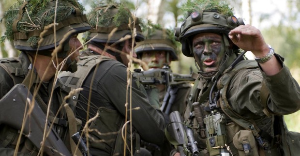 Soldaten des österreichischen Bundesheeres in voller Ausrüstung während einer Übung im Wald. Die Gesichter sind mit Tarnfarbe bemalt, Helme mit Zweigen getarnt. Ein Soldat gibt gestikulierend Anweisungen