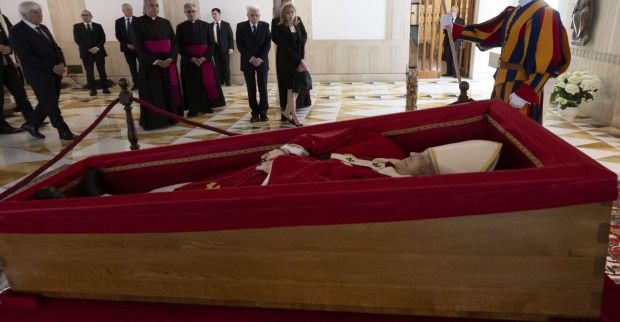 Papst Franziskus liegt im offenen Sarg in rotem liturgischem Gewand. Um den Sarg versammeln sich Geistliche, Politiker und ein Mitglied der Schweizergarde.