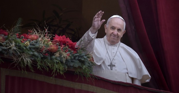 Papst Franziskus steht in weißem Gewand auf einem festlich geschmückten Balkon, winkt lächelnd in die Menge – im Hintergrund roter Vorhang und Tannenschmuck.
