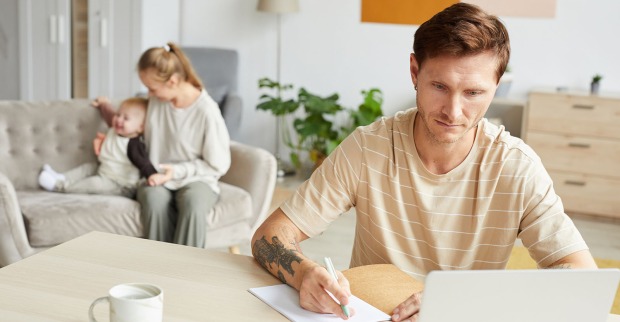 Junger Vater sitzt am Laptop am Küchentisch, während im Hintergrund seine Partnerin mit einem Kleinkind auf dem Sofa spielt
