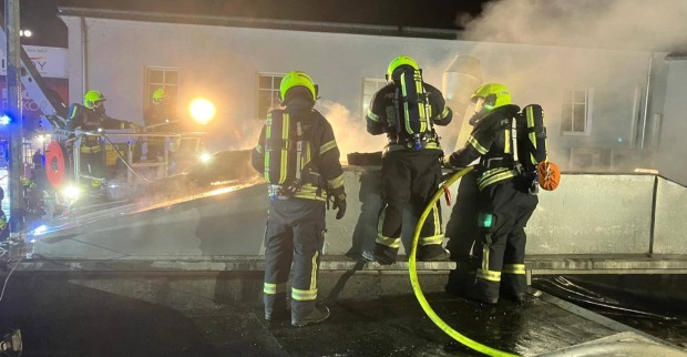 Vier Feuerwehrleute mit Atemschutzgeräten stehen nachts auf einem Flachdach im Einsatz gegen einen Brand; dichter Rauch steigt auf, ein Hubsteiger ist im Hintergrund im Einsatz.