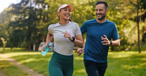 Eine Frau und ein Mann joggen. Beide lachen. Sie hat eine grüne Hose und ein weißes T-Shirt an, zudem trägt sie eine Schirmkappe. In einer Hand hält sie eine grüne Trinkflasche. Der Mann mit Vollbart hat eine blaues T-Shirt und eine schwarze Jogging-Hose an. Im Hintergrund sind Bäume und eine Wiese zu sehen.