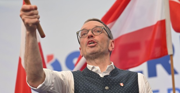 Herbert Kickl hält eine Fahne in die Höhe und spricht energisch auf der Bühne, im Hintergrund rot-weiß-rote österreichische Fahnen.