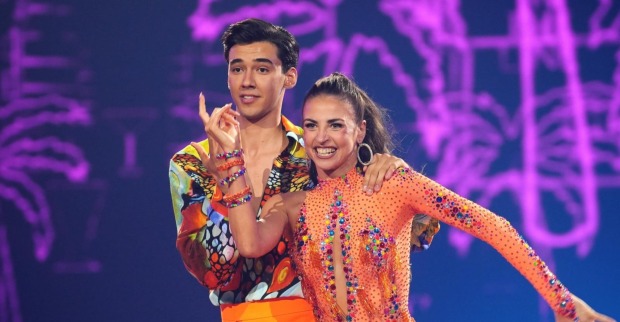 Diego Pooth und seine Tanzpartnerin Ekaterina Leonova lächeln auf der "Let's Dance"-Bühne im Finale 2025. Beide tragen bunte, glitzernde Kostüme, die Kulisse ist violett beleuchtet.