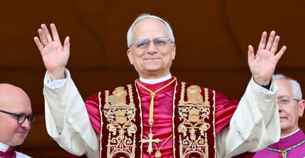 Papst Leo XIV. steht in päpstlicher liturgischer Kleidung auf dem Balkon des Petersdoms und hebt beide Hände zum Gruß. Links und rechts neben ihm stehen Kirchenvertreter.