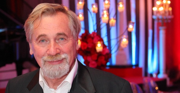 Porträt von Peter Rapp mit weißem Haar und Bart, freundlich lächelnd in festlichem Ambiente mit Kerzenlichtern und rotem Blumenschmuck im Hintergrund.