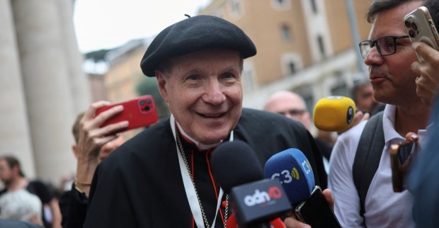 Kardinal Christoph Schönborn, freundlich lächelnd mit schwarzem Barett und rotem Kardinalsrang, wird von mehreren Journalisten mit Mikrofonen und Smartphones umringt. Im Hintergrund sind Menschen und Gebäude in Rom zu sehen.