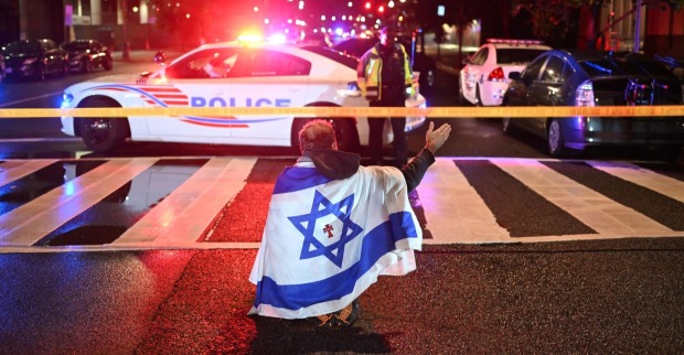 Ein Mann mit einer israelischen Flagge auf dem Rücken kniet nachts auf einer gesperrten Straße vor einem mit Blaulicht beleuchteten Polizeiwagen in Washington D.C. – Tatort eines tödlichen Anschlags auf israelische Botschaftsmitarbeiter.