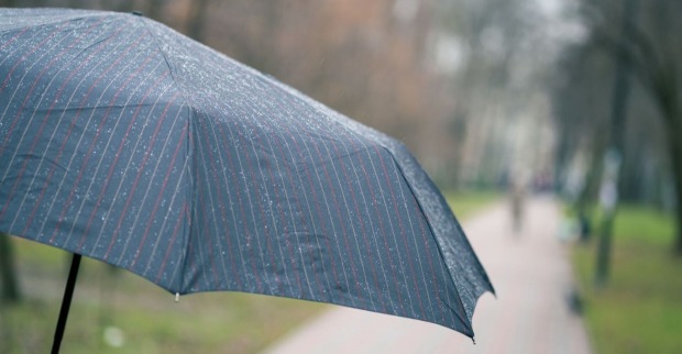 Ein Regenschirm mit feinen Regentropfen steht geöffnet auf einem Gehweg in einem Park; der Hintergrund ist unscharf mit Bäumen und Fußgängern.