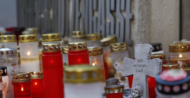 Zahlreiche rote und weiße Grablichter stehen vor einer Wand mit dem Schriftzug „Dreierschützengasse“. Im Vordergrund ein graues Steinkreuz mit der Inschrift „Wir vermissen Dich“ und einer Engelsfigur.