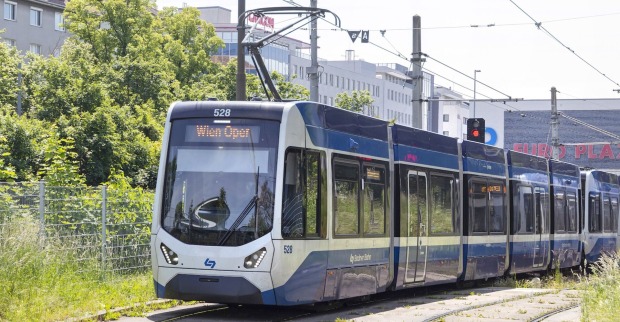 Moderne Garnitur der Badner Bahn mit Zielanzeige „Wien Oper“, fährt durch eine begrünte Gleisanlage vor städtischem Hintergrund, aufgenommen bei Tageslicht.