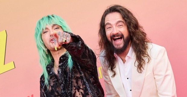 Bill Kaulitz mit neongrüner Frisur und durchsichtiger schwarzer Bluse zeigt lachend auf die Kamera, neben ihm steht Tom Kaulitz in weißem Anzug, beide posieren fröhlich vor rotem Hintergrund.