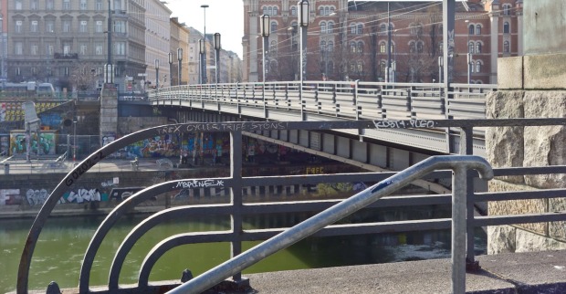 Blick auf die Augartenbrücke in Wien mit Blick auf den Donaukanal und Graffiti an den Ufermauern; im Vordergrund ein Geländer mit Schmierereien, im Hintergrund historische Gebäude.