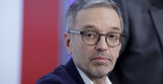 Herbert Kickl mit ernstem Gesichtsausdruck, trägt Brille und Sakko, vor rotem und weißem Hintergrund.