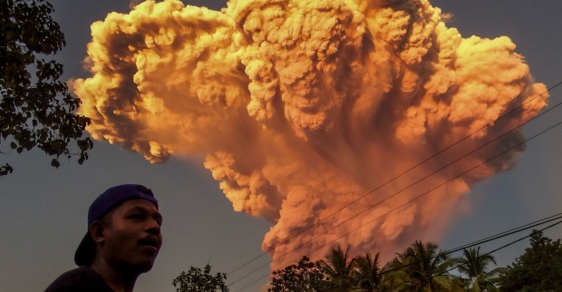 Ein Mann steht im Vordergrund, während im Hintergrund eine gewaltige orangefarbene Aschewolke vom ausbrechenden Vulkan Lewotobi Laki-Laki auf der indonesischen Insel Flores aufsteigt.
