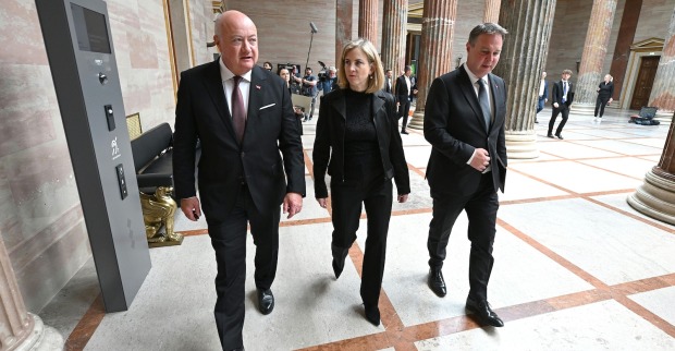 Bundeskanzler Christian Stocker, Außenministerin Beate Meinl-Reisinger und Vizekanzler Andreas Babler – gehen durch die Säulenhalle des Parlamentsgebäudes in Wien, begleitet von Medienvertretern.