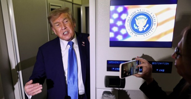 Donald Trump spricht in einem Flugzeug mit Medienvertretern, neben ihm ein Bildschirm mit dem Präsidentensiegel der USA. Eine Person filmt ihn mit dem Smartphone.