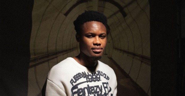 Kelvin Boateng vor einer Leinwand sitzend, welche den Durchgang der U-Bahn-Station Altes Landgut zeigt