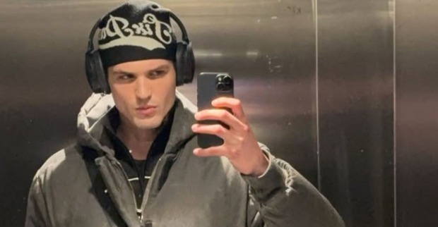 Pierre trägt grau-schwarz-weiße Kleidung, eine Jacke, eine Mütze und Kopfhörer und macht ein Spiegel-Selfie