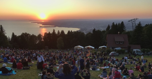 Menschen sitzen auf einer Wiese am Pfänder und blicken auf den Bodensee bei Sonnenuntergang während des Sonnwend-Fests mit Feuerstelle und Festzelten.