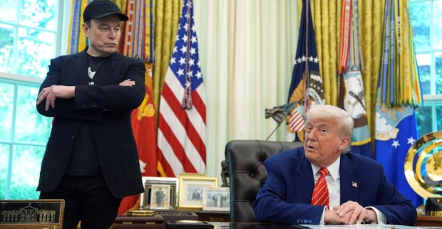 Elon Musk steht mit verschränkten Armen neben Donald Trump, der am Schreibtisch im Oval Office sitzt. Im Hintergrund US-Flaggen und militärische Standarten.