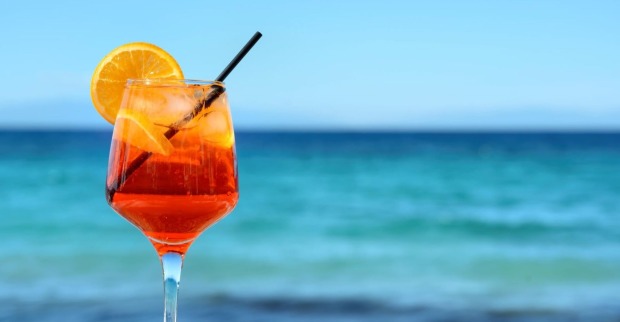 Ein Glas Aperol Spritz mit Orangenscheibe und Strohhalm vor türkisblauem Meer und klarem Himmel – Symbol für Sommerurlaub und Urlaubsstimmung