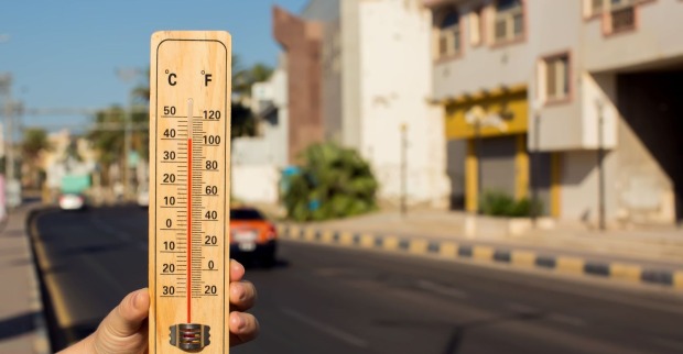 Hand hält Thermometer, das über 40 Grad Celsius zeigt, vor einer sonnigen Straße mit Gebäuden und Autos.