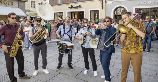 Sechs Musiker der Band Sloerfcats spielen mit Saxophon, Sousaphon, Trommel, Banjo, Trompete und Posaune in einer belebten Salzburger Altstadtgasse, umringt von einem großen Publikum.