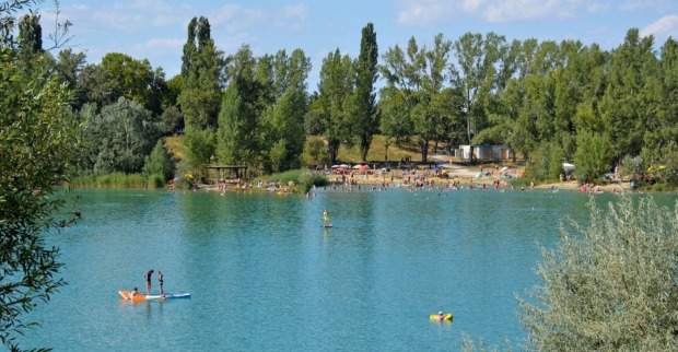 Badeteich Hirschstetten in Wien an einem sonnigen Tag, viele Menschen schwimmen und entspannen am Ufer, im Vordergrund Kinder auf einem Stand-Up-Paddle.