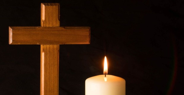 Eine leuchtende weiße Kerze mit einem Holzkreuz vor schwarzem Hintergrund als Zeichen für Trauer