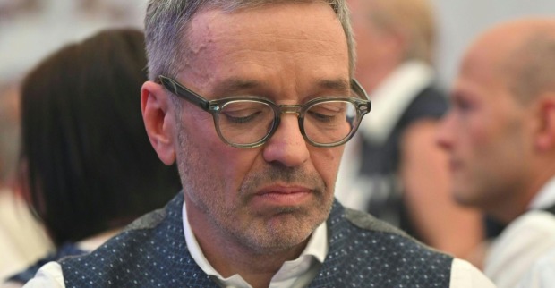 Herbert Kickl mit gesenktem Blick und ernstem Gesichtsausdruck, trägt Brille und Trachtenweste; im Hintergrund unscharf andere Personen.