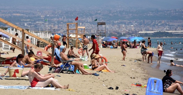 Menschen an einem überfüllten Sandstrand in Catania, Sizilien, bei extremer Sommerhitze; viele sitzen unter Sonnenschirmen oder gehen ins Meer zur Abkühlung.