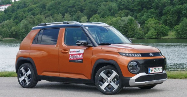 Ein kaffeebrauner Hyundai Inster parkt am Ufer eines Flusses. Am anderen Ufer ist ein dichter Laubwald zu sehen.